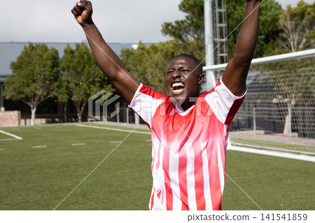 African American male athlete celebrating goal, raising arms on turf field in front of goalpost net 141541859