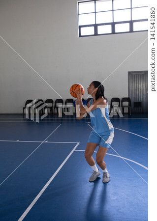 African American woman holding orange basketball shooting on blue court near black chairs African American woman holding orange basketball shooting on blue court near black chairs 141541860