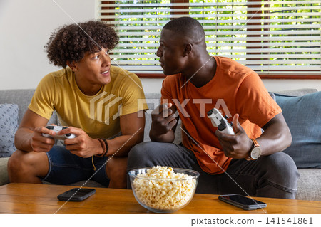 Diverse male friends playing console games holding controllers on sofa at home, popcorn bowl Diverse male friends playing console games holding controllers on sofa at home, popcorn bowl 141541861
