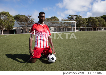 African American male soccer player kneeling on field by goalpost resting on ball in red jersey African American male soccer player kneeling on field by goalpost resting on ball in red jersey 141541863