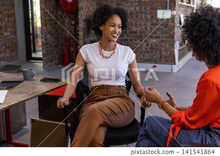 Diverse female coworkers sitting at desk chairs talking in office with coffee mug 141541864