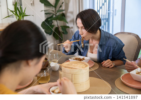Diverse friends sitting at dining table in home eating noodles from steamer with chopsticks Diverse friends sitting at dining table in home eating noodles from steamer with chopsticks 141541897