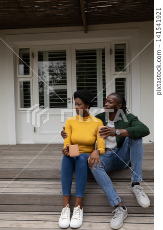 African american couple sitting on wooden porch steps under slatted pergola with ceramic mugs 141541921