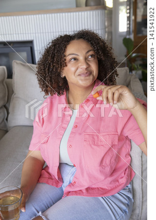 African American woman in casual outfit snacking on cookie, holding glass on couch at home African American woman in casual outfit snacking on cookie, holding glass on couch at home 141541922