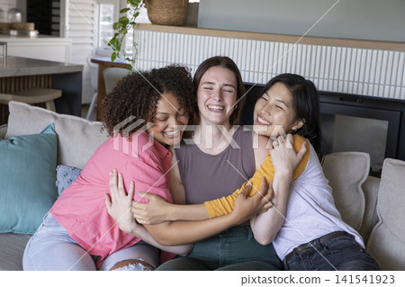Diverse female friends hugging on sofa in home living room, laughing with fireplace backdrop Diverse female friends hugging on sofa in home living room, laughing with fireplace backdrop 141541923