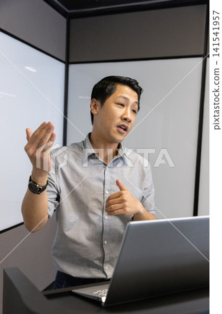 Asian man presenting behind podium in meeting room with laptop computer, frosted glass panels Asian man presenting behind podium in meeting room with laptop computer, frosted glass panels 141541957
