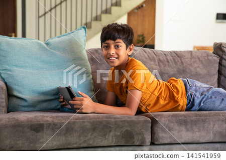 Indian boy lying on gray sofa in living room holding handheld gaming device near turquoise cushion Indian boy lying on gray sofa in living room holding handheld gaming device near turquoise cushion 141541959