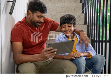 Indian father and son sitting on staircase landing at home and viewing tablet computer together Indian father and son sitting on staircase landing at home and viewing tablet computer together 141541966