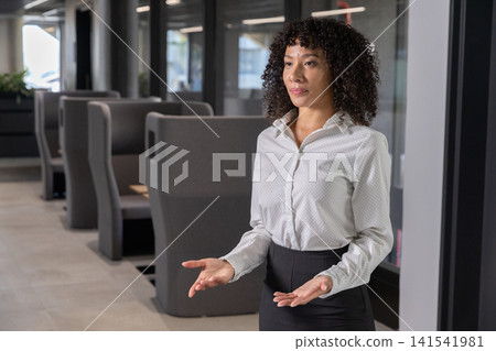 African american woman standing by seating booths, plant, glass walls and lights in office African american woman standing by seating booths, plant, glass walls and lights in office 141541981