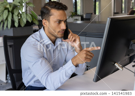 Asian man talking on smartphone while pointing at computer monitor on modern office desk Asian man talking on smartphone while pointing at computer monitor on modern office desk 141541983
