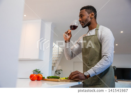 African american man wearing apron sniffing red wine at kitchen island with sliced zucchini African american man wearing apron sniffing red wine at kitchen island with sliced zucchini 141541987