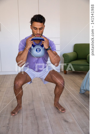 Man with 8 kg kettlebell close to chest while squatting in living room workout area Man with 8 kg kettlebell close to chest while squatting in living room workout area 141541988