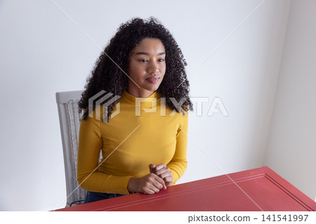 African american woman sitting at red table on chair and gazing off-camera in home workspace African american woman sitting at red table on chair and gazing off-camera in home workspace 141541997