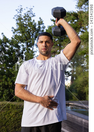 Man wearing white t-shirt and shorts holding black hexagonal dumbbell overhead on wooden deck Man wearing white t-shirt and shorts holding black hexagonal dumbbell overhead on wooden deck 141541998