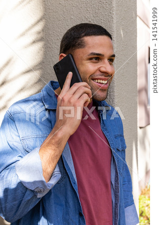 Hispanic man in denim shirt leaning near wall outside phoning on smartphone amid leaf shadows Hispanic man in denim shirt leaning near wall outside phoning on smartphone amid leaf shadows 141541999