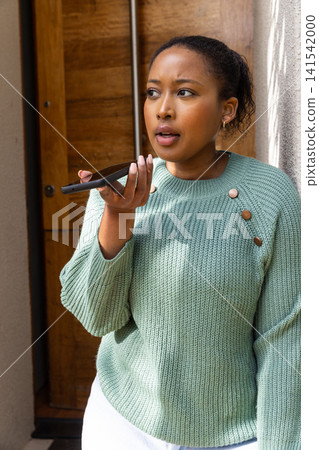 African american woman wearing sweater at entryway holding smartphone near mouth, speaking African american woman wearing sweater at entryway holding smartphone near mouth, speaking 141542000