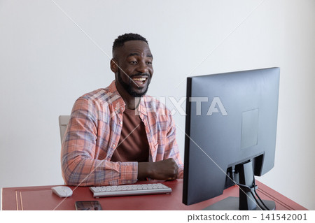 African american man typing on wireless keyboard, smiling at computer monitor in home office 141542001