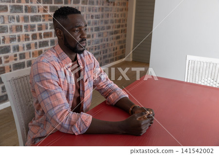 African american man sitting on white wicker chair against exposed brick wall gazing at table 141542002