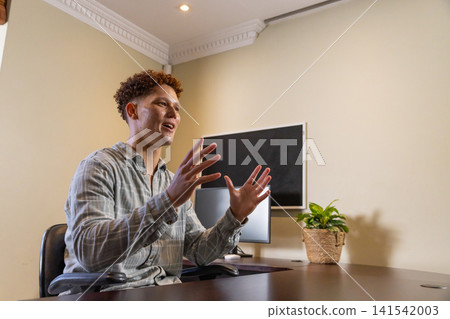 Man gesturing while speaking at wooden desk in office with monitor, plant, copy space Man gesturing while speaking at wooden desk in office with monitor, plant, copy space 141542003