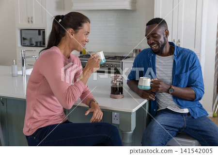 Diverse couple holding coffee mugs and smiling at each other at kitchen with french press 141542005