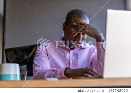 African american man rubbing forehead, sitting at desk in office with laptop monitor African american man rubbing forehead, sitting at desk in office with laptop monitor 141542013