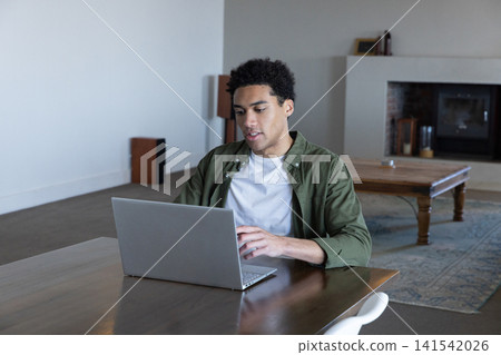 African american man typing on silver laptop while sitting at wooden table in living area African american man typing on silver laptop while sitting at wooden table in living area 141542026