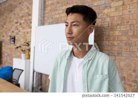 Asian man standing and gazing off to right in office with whiteboard beanbag, copy space Asian man standing and gazing off to right in office with whiteboard beanbag, copy space 141542027
