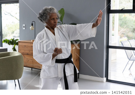 Senior african american woman wearing white gi, black belt practicing karate in living room 141542031