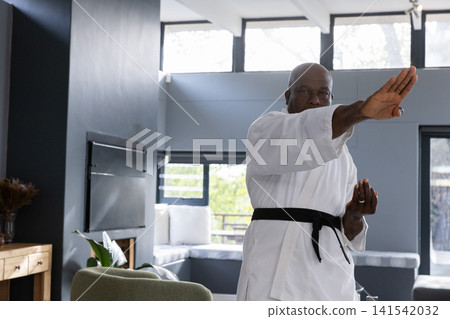 Senior african american man wearing karate gi, belt punching in living room by bench Senior african american man wearing karate gi, belt punching in living room by bench 141542032