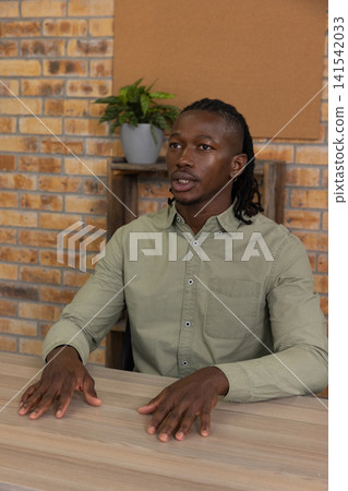 African american man sitting at table in office with shelf and holding potted plant and cork board African american man sitting at table in office with shelf and holding potted plant and cork board 141542033