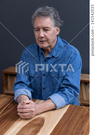 Senior man wearing denim shirt sitting at wooden table clasping hands showing wedding ring Senior man wearing denim shirt sitting at wooden table clasping hands showing wedding ring 141542035