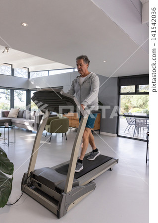 Senior man walking on treadmill at home wearing sportswear with sliding glass doors, armchair Senior man walking on treadmill at home wearing sportswear with sliding glass doors, armchair 141542036