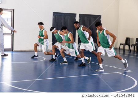 Diverse male basketball team in jerseys practicing lunges on court under coach instruction Diverse male basketball team in jerseys practicing lunges on court under coach instruction 141542041