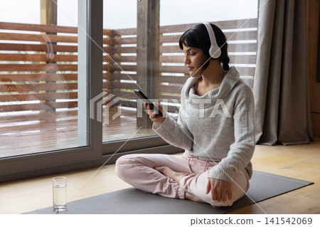 Asian adult woman sitting on yoga mat by glass door wearing headphones holding smartphone Asian adult woman sitting on yoga mat by glass door wearing headphones holding smartphone 141542069