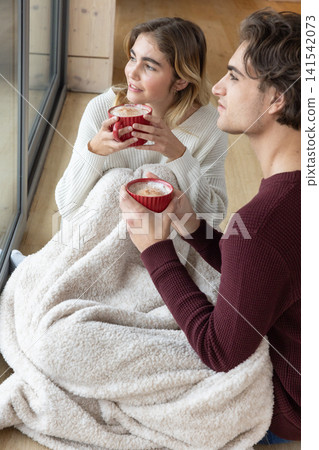 Couple sitting on wooden floor next to large window wrapped in sherpa blanket holding red mugs Couple sitting on wooden floor next to large window wrapped in sherpa blanket holding red mugs 141542073