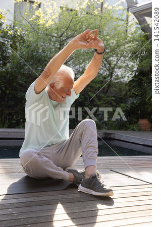 Senior man stretching arms overhead on black yoga mat on poolside wooden deck with garden foliage Senior man stretching arms overhead on black yoga mat on poolside wooden deck with garden foliage 141542089