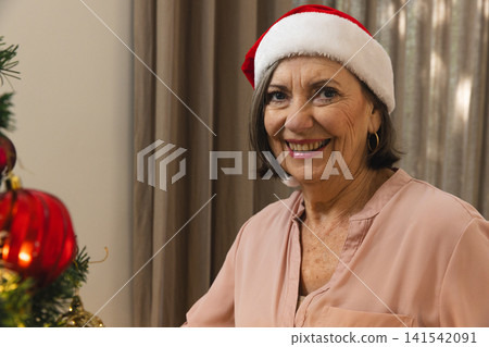 Senior woman wearing Santa hat, pink blouse sitting by Christmas tree in living room, copy space Senior woman wearing Santa hat, pink blouse sitting by Christmas tree in living room, copy space 141542091