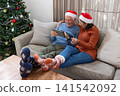 Senior couple sitting at home wearing Santa hats browsing tablet with credit card by Christmas tree 141542092
