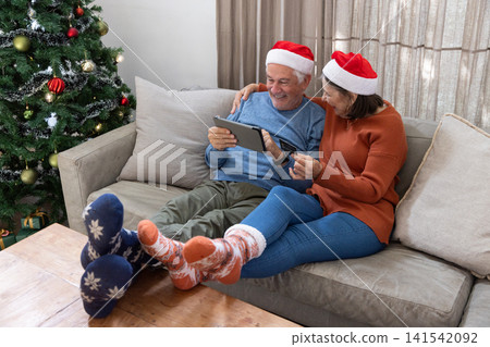 Senior couple sitting at home wearing Santa hats browsing tablet with credit card by Christmas tree Senior couple sitting at home wearing Santa hats browsing tablet with credit card by Christmas tree 141542092