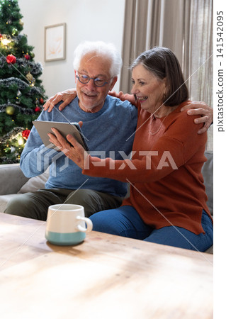 Senior couple sharing tablet while sitting on sofa in living room near Christmas tree and mug Senior couple sharing tablet while sitting on sofa in living room near Christmas tree and mug 141542095