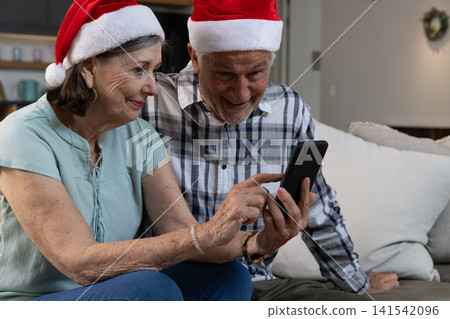 Senior couple sitting on gray sofa in living room pointing at smartphone wearing Santa hats Senior couple sitting on gray sofa in living room pointing at smartphone wearing Santa hats 141542096