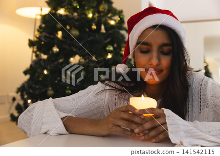 Asian woman wearing Santa hat in living room holding lit candle by Christmas tree, copy space 141542115
