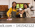 Diverse mother and daughter wearing aprons wiping countertop with cloth and spray bottle in kitchen 141542122