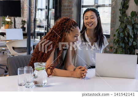 Diverse female coworkers collaborating over laptop at table in office with pitcher of lime water Diverse female coworkers collaborating over laptop at table in office with pitcher of lime water 141542141