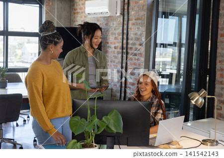 Diverse female coworkers collaborating around computer monitor at office with tablet, potted plant Diverse female coworkers collaborating around computer monitor at office with tablet, potted plant 141542143