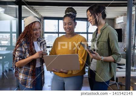 Diverse female colleagues collaborating around laptop and tablet in modern loft office with skyline Diverse female colleagues collaborating around laptop and tablet in modern loft office with skyline 141542145