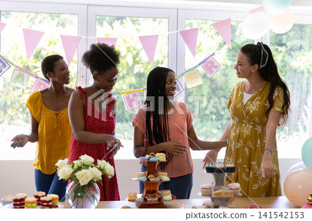 Diverse female friends gathering around table at home with tiered cupcakes and pink pennant banners Diverse female friends gathering around table at home with tiered cupcakes and pink pennant banners 141542153