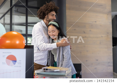 Diverse coworkers hugging beside office desk in casual wear with monitor showing charts, balloon Diverse coworkers hugging beside office desk in casual wear with monitor showing charts, balloon 141542154