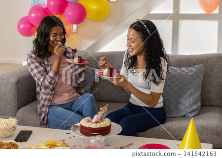Diverse mom and teenage daughter eating birthday red velvet cake on sofa in living room with plates Diverse mom and teenage daughter eating birthday red velvet cake on sofa in living room with plates 141542163