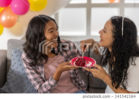Diverse mom and teenage daughter laughing while sharing cake on red plate on sofa in living room Diverse mom and teenage daughter laughing while sharing cake on red plate on sofa in living room 141542164